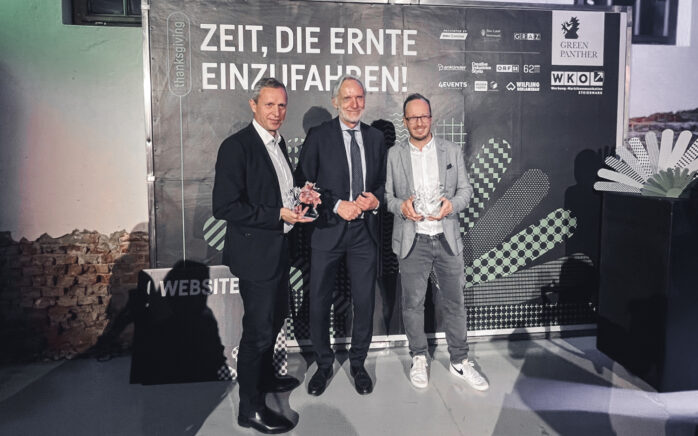 Das Bild zeigt drei Männer, die lächelnd für Fotos posieren. Zwei von ihnen halten einen Award in der Hand.