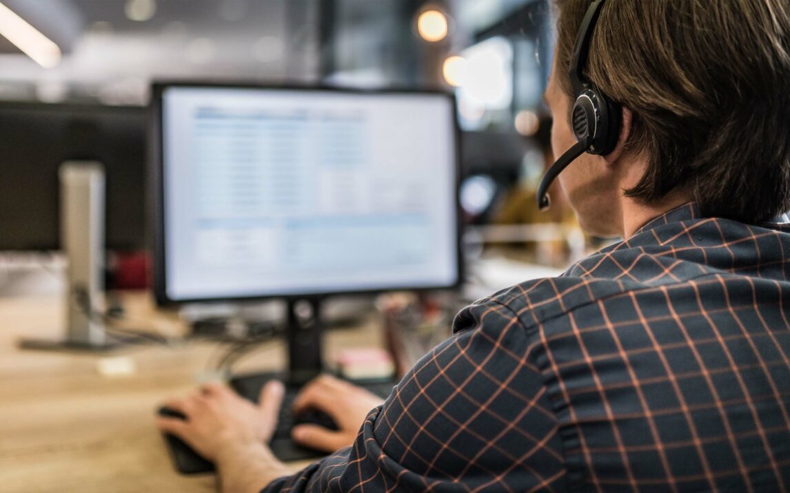Over-the shoulder view of a man with a headset with a microphone working in a call center. Person wearing a headset works on a computer in an office, facing the screen, which displays data.