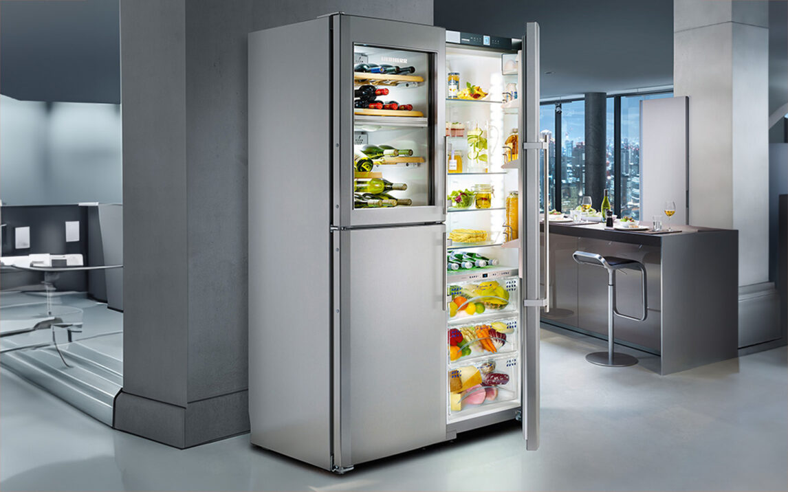 Header-reference-liebherr-hausgeräte-EN An open LIEBHERR stainless steel refrigerator in a modern kitchen, showcasing assorted fruits, vegetables, bottles of drinks, and various groceries on multiple shelves.