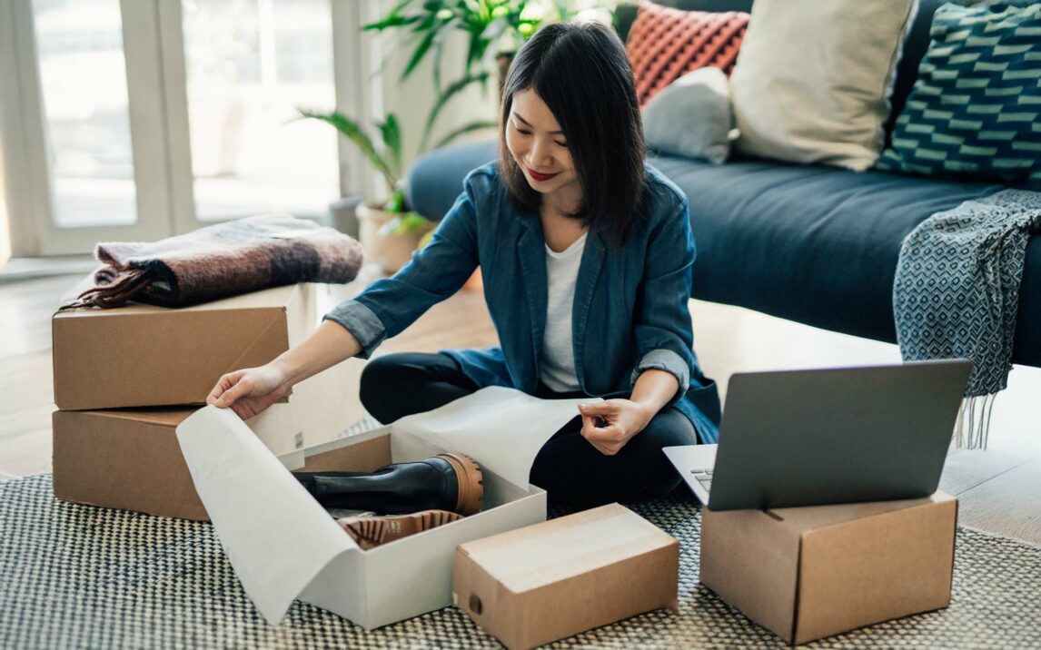 Young woman using laptop and packing packages, fulfilling online orders. E-commerce and online shopping helps small business. Working from home concept. Young woman using laptop and packing packages, fulfilling online orders. E-commerce and online shopping helps small business. Working from home concept.