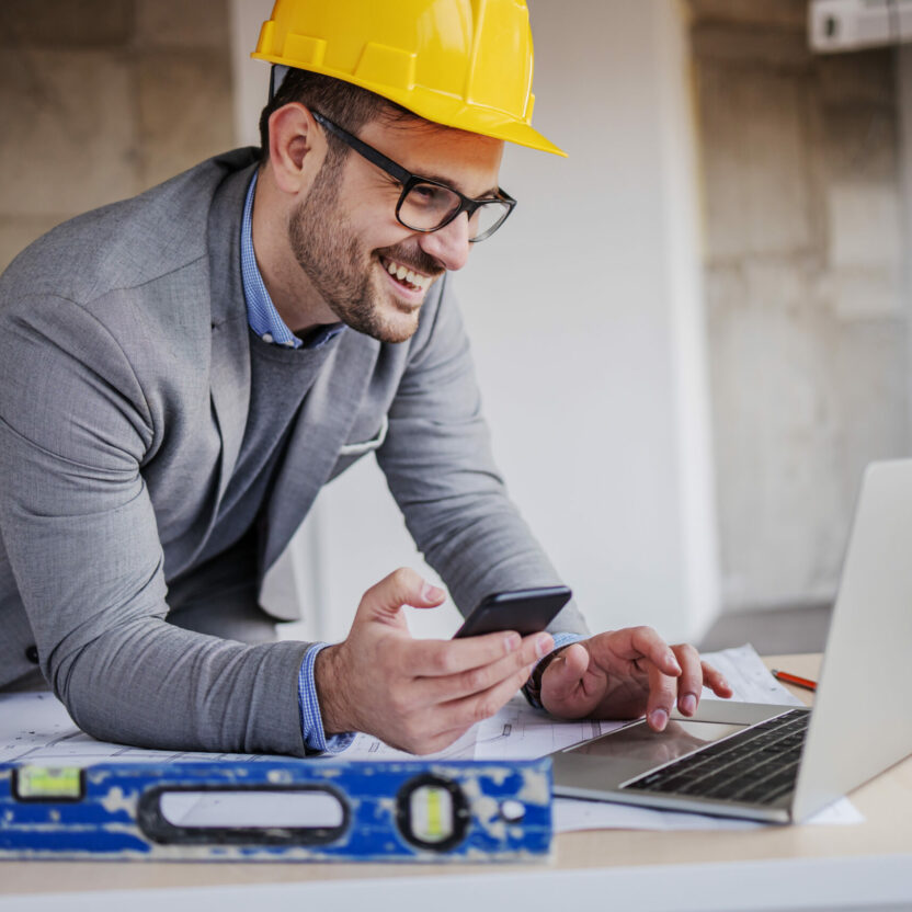 Young smiling architect in suit with helmet on head holding smart phone and using laptop while leaning on table in building in construction process. Architekt auf Baustelle