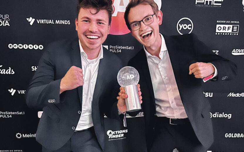Zwei junge Männer in Anzügen feiern begeistert und halten gemeinsam einen gläsernen Award in die Kamera. Im Hintergrund ist eine Sponsorenwand mit Logos zu sehen.
