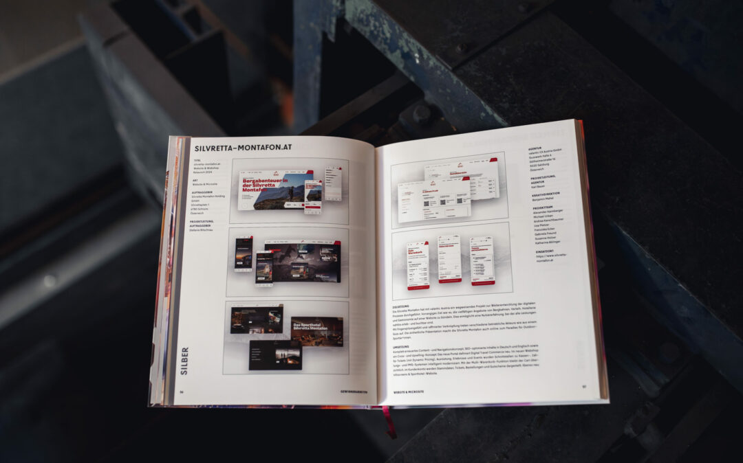 Auf einer dunklen Oberfläche liegt ein offenes Buch, dessen Seiten Website-Design-Layouts und Einblicke in das Projekt Silver Delta Montafon offenbaren, wie es im AMMA Jahrbuch 2025 vorgestellt wird.