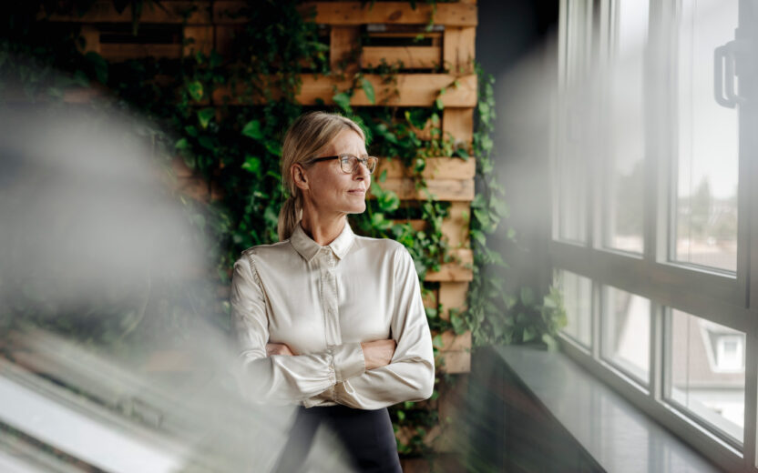 A woman with glasses stands indoors, arms crossed, contemplating agile Produktionsplanung as she gazes out a window. A wooden wall with green plants behind her adds a touch of nature to the otherwise serene scene.