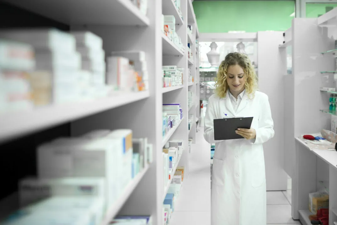Anaplan | Biotech Vertriebsplanung mit Anaplan Biotech Vertriebsplanung mit Anaplan Frau steht mit weißem Kittel in einem Raum mit Medikamenten in den Regalen und notiert etwas auf einem Klemmbrett