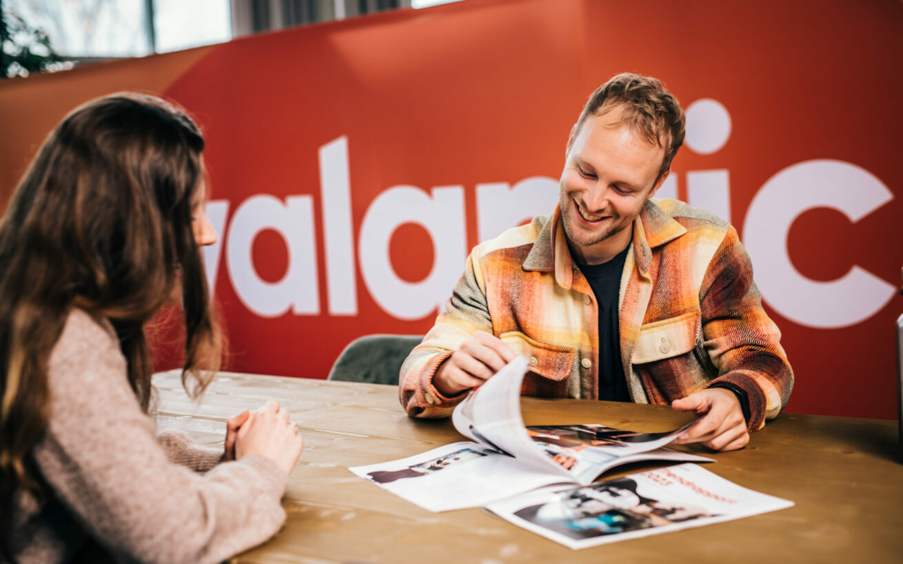 Valantic_SetVexy_0A1A8260 Twee mensen zitten aan een tafel, de een laat de ander een tijdschrift zien. Beiden zijn in gesprek, met een rode muur en een gedeeltelijke tekst "alamic" op de achtergrond.