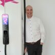 Eine Person im weißen Hemd steht neben einer Ladestation für Elektrofahrzeuge mit digitaler Anzeige und lächelt in die Kamera.