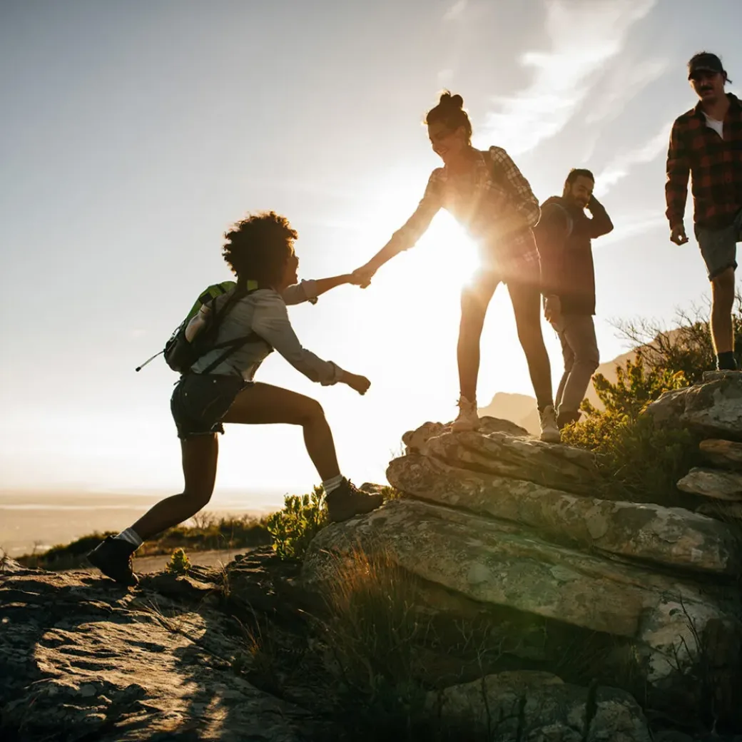 people-on-mountain-hike-at-sunset-649871644 Group of hikers on a mountain. Woman helping her friend to climb a rock. Young people on mountain hike at sunset.