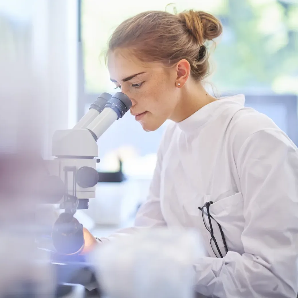 research-student-scientist-542576580 A female research scientist is analysing a sample on her microscope in a microbiology lab . The lab is brightly lit with natural light. Blurred glassware at side of frame provides copy space