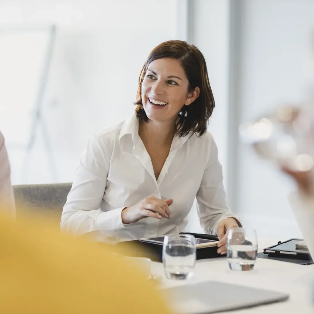 woman-smiling-at-business-meeting-1200927481 Businesswoman smiling at meeting table, listening, learning, success, happiness